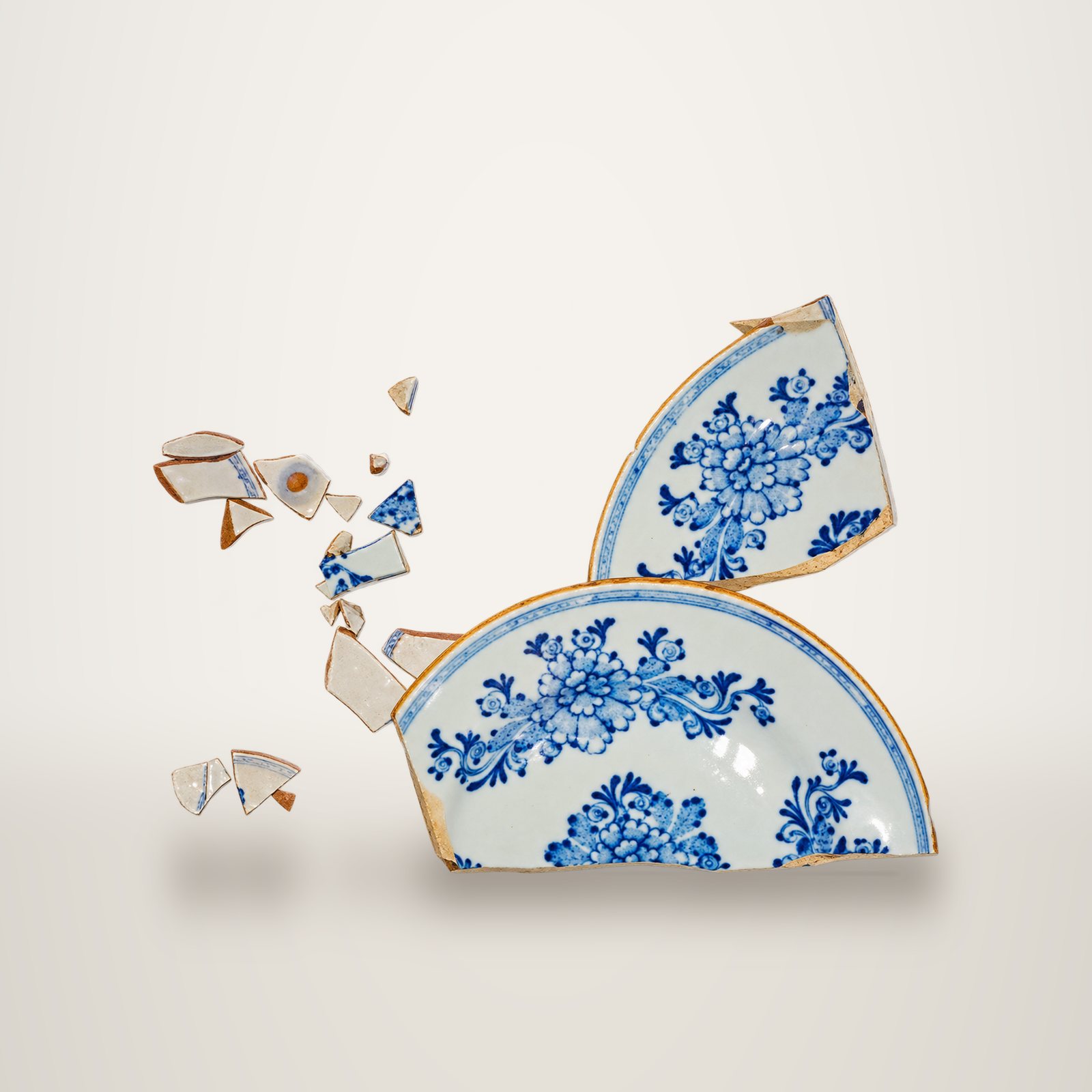 Une assiette ancienne en faïence à décor floral bleu sur fond blanc, brisée en plusieurs grands morceaux et petits éclats, photographiée sur fond blanc uni.