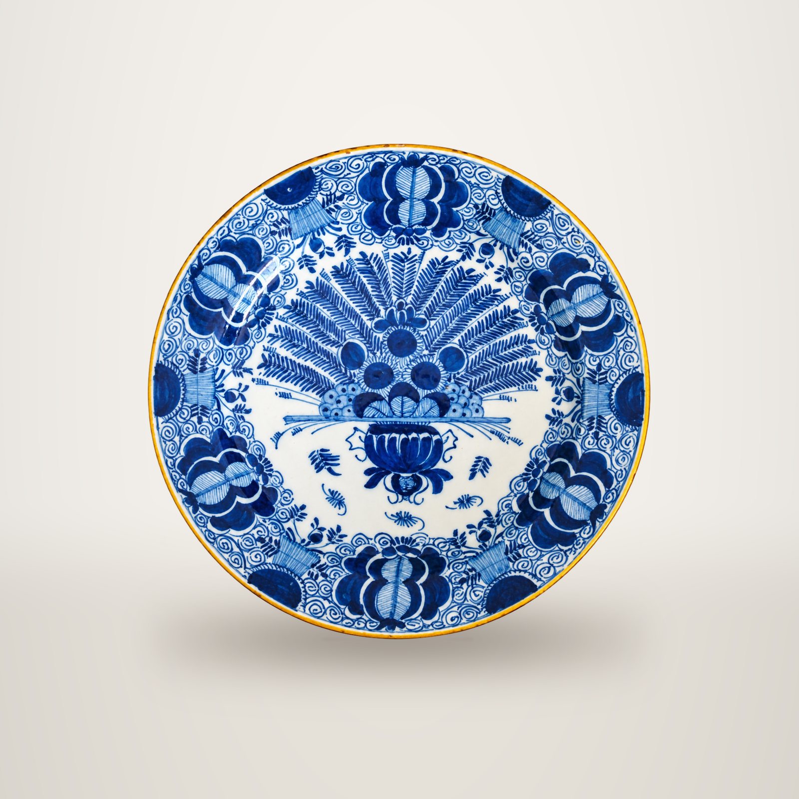 Assiette ancienne en faïence de Delft à motifs bleus et blancs, avec un bouquet de plumes de paon au centre et des fleurs stylisées sur le bord.