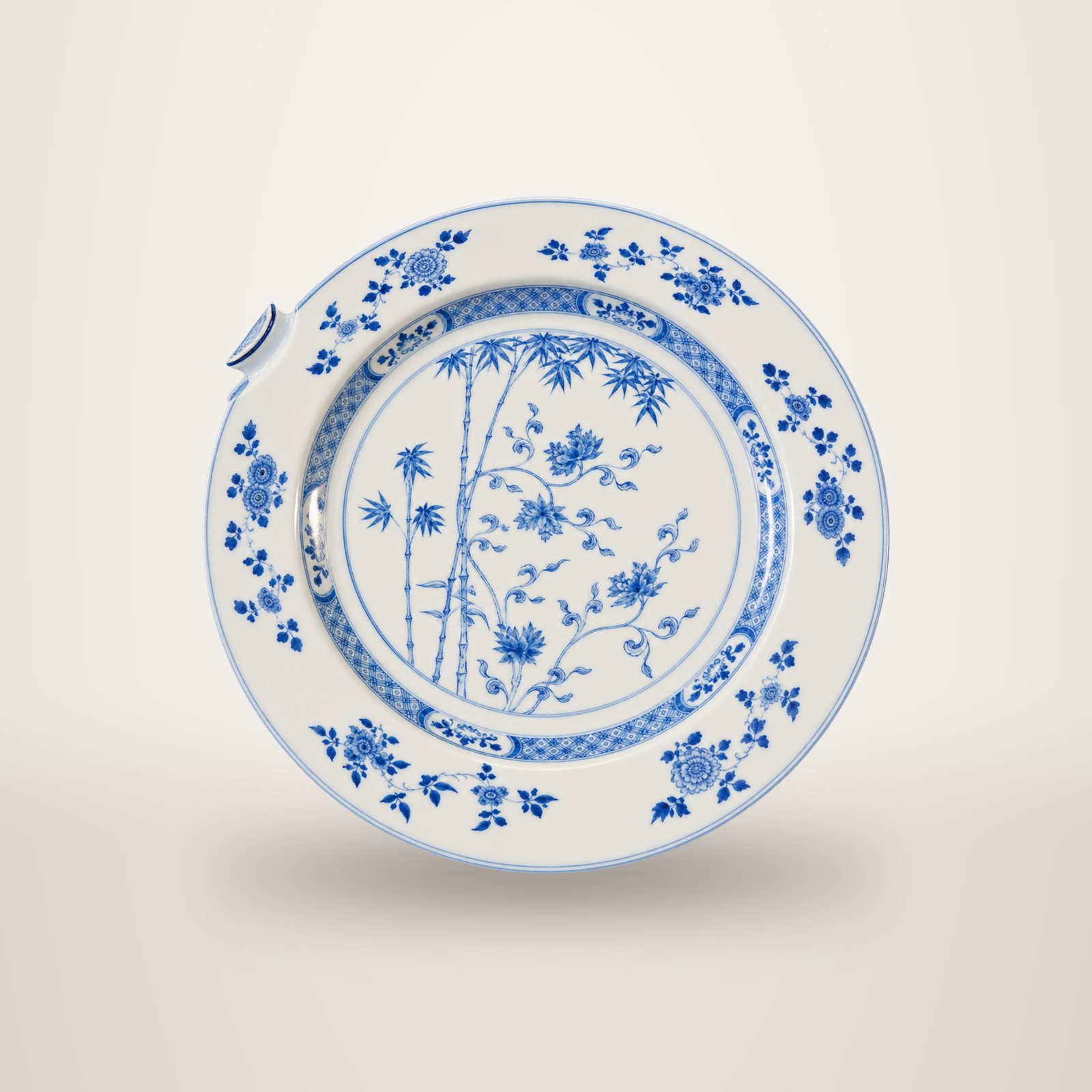 Une assiette ancienne en porcelaine blanche ornée de bambous et de motifs floraux bleus, avec un petit éclat sur le bord, photographiée sur fond blanc.