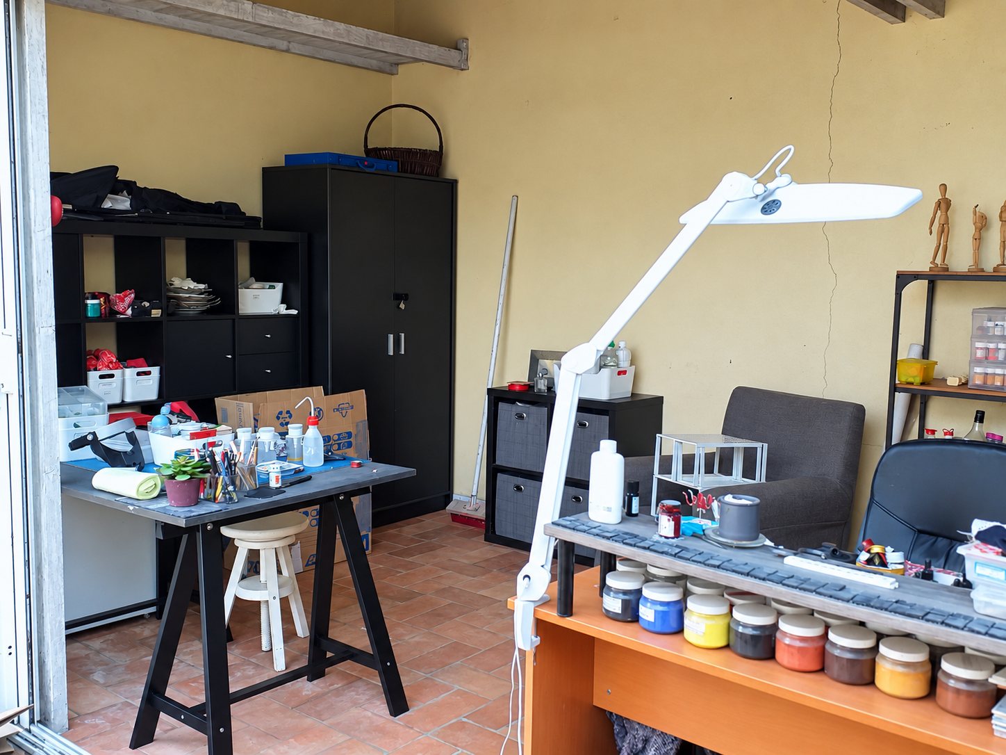 Intérieur d'un atelier au sol en terre cuite, avec une table de travail couverte de pinceaux, une grande lampe blanche articulée et des pots de pigments colorés alignés sur un bureau.