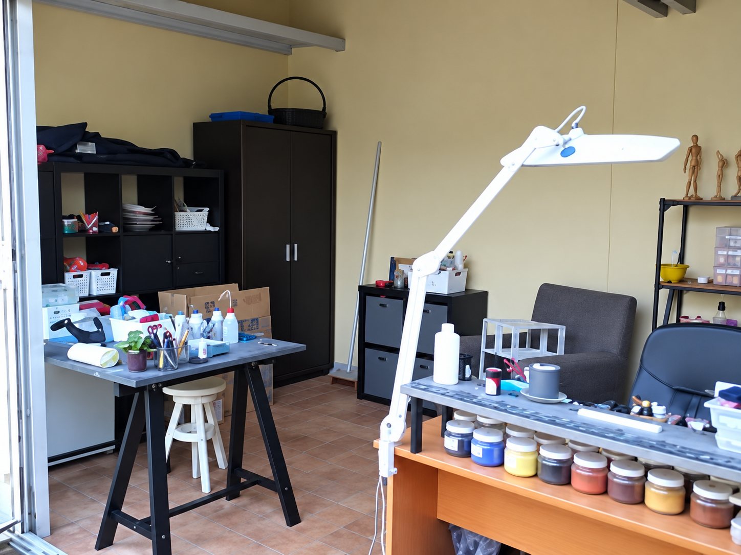 Intérieur d'un atelier avec un établi, une lampe articulée blanche, des pots de peinture alignés et des étagères de rangement au fond.