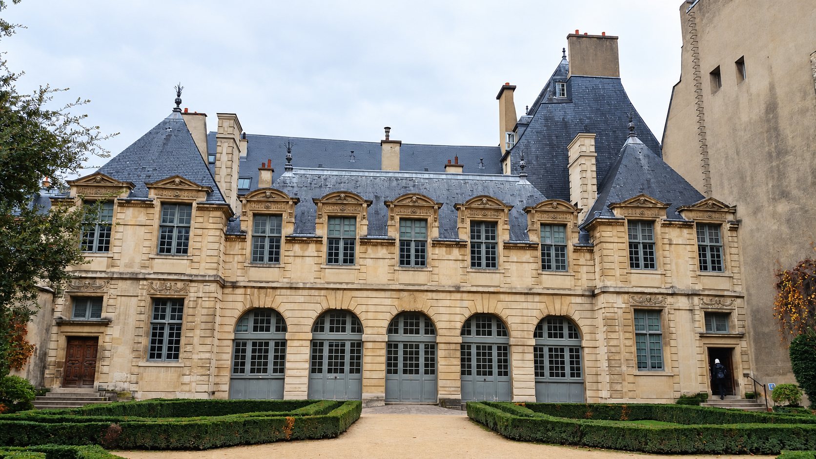 Façade en pierre claire d'un hôtel particulier classique aux toits d'ardoise, précédée d'un jardin à la française avec haies de buis taillées.