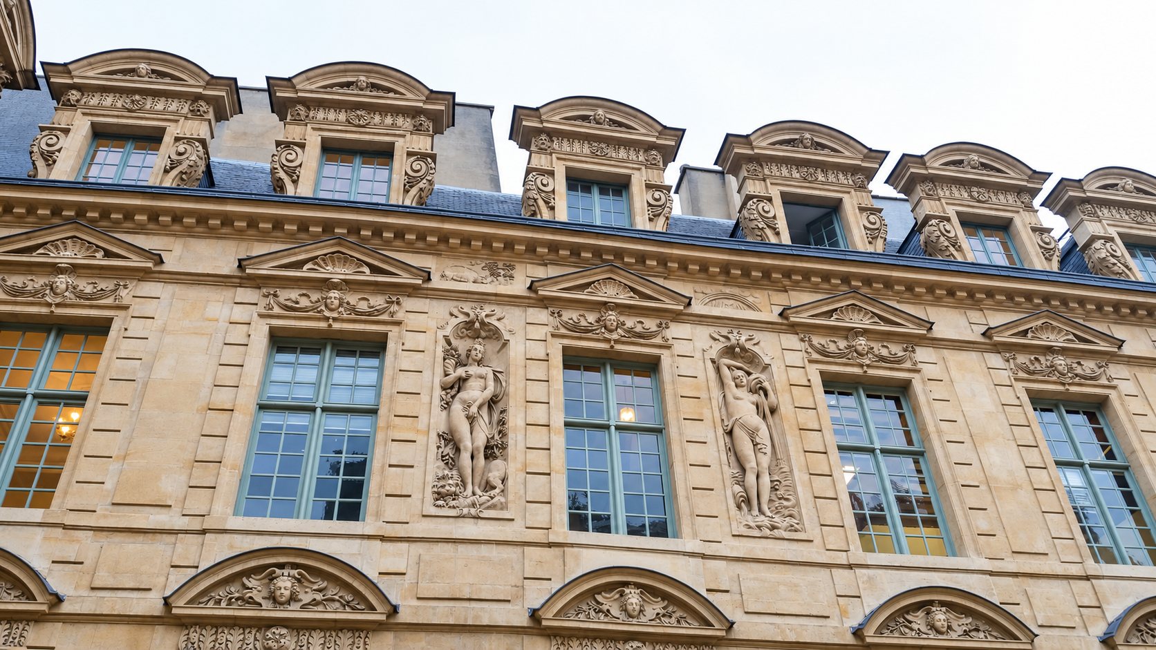 Façade d'un bâtiment ancien en pierre claire, ornée de statues, de frontons sculptés et de lucarnes décorées sous un ciel gris.