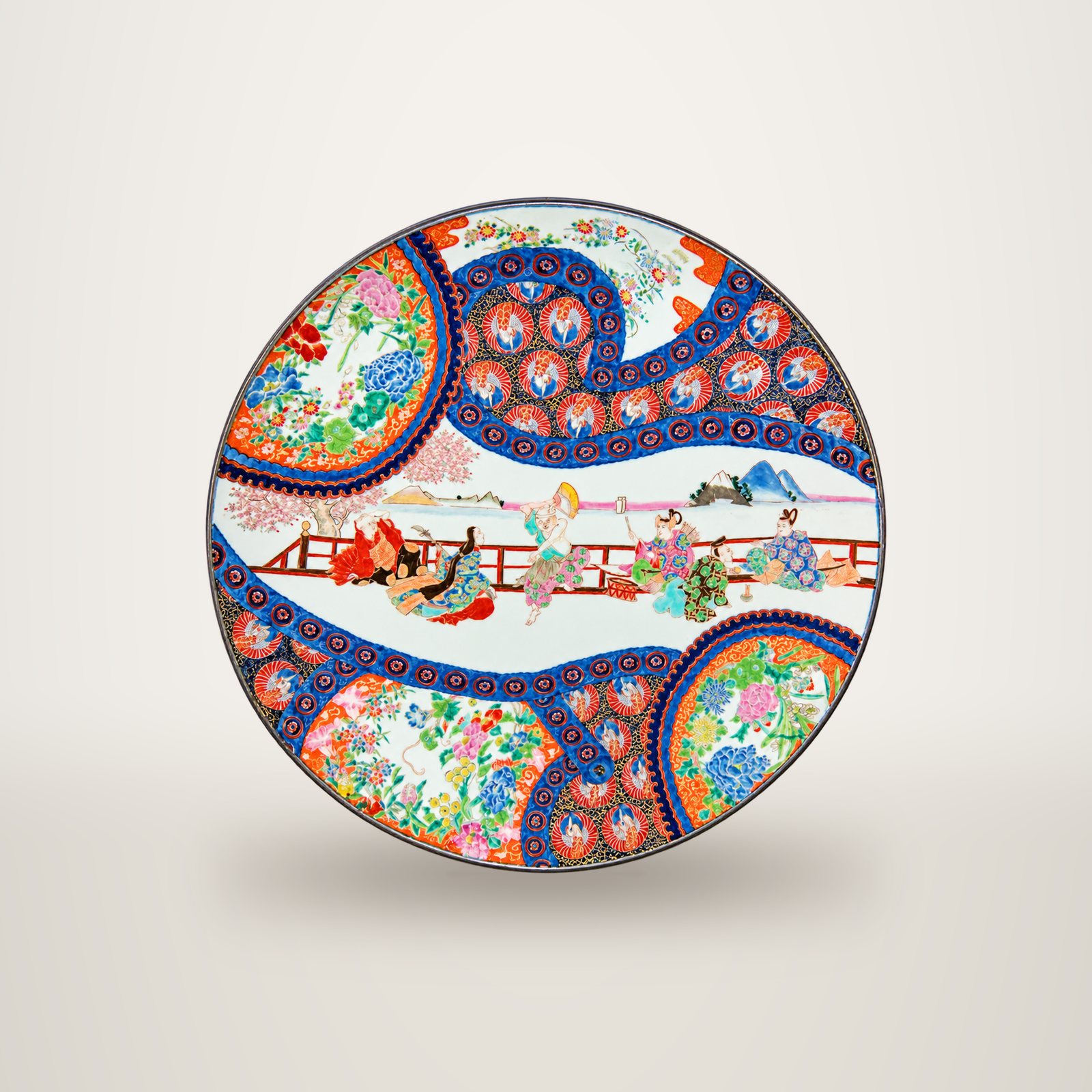 Grand plat rond en porcelaine japonaise orné de personnages près d'une balustrade, de motifs floraux colorés et de bordures bleues et rouges.