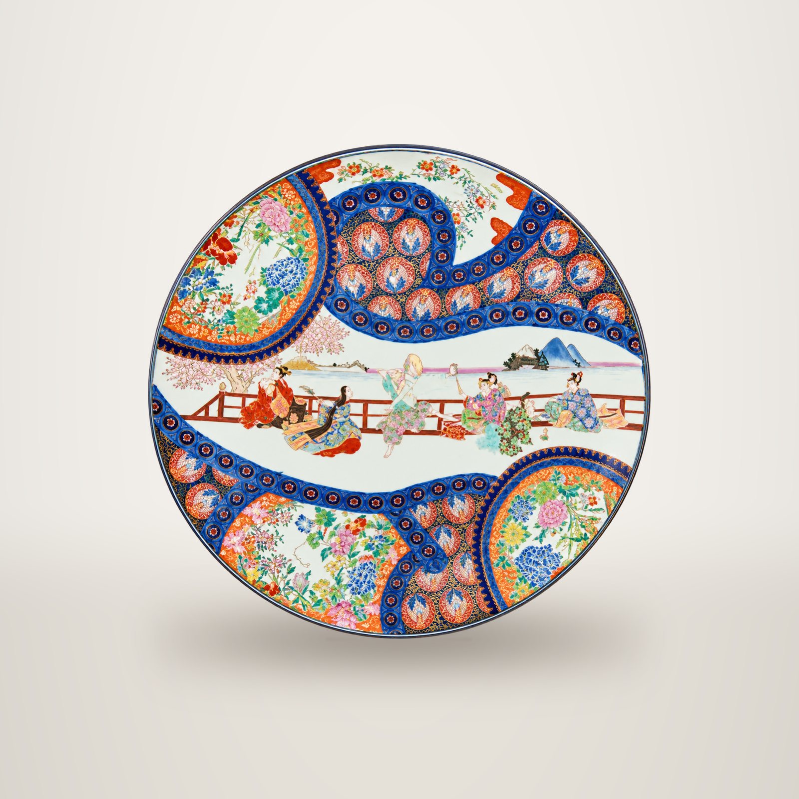 Grand plat en porcelaine aux motifs japonais colorés, avec des personnages près d'une balustrade et des médaillons de fleurs bleus, rouges et verts, présenté sur fond blanc.