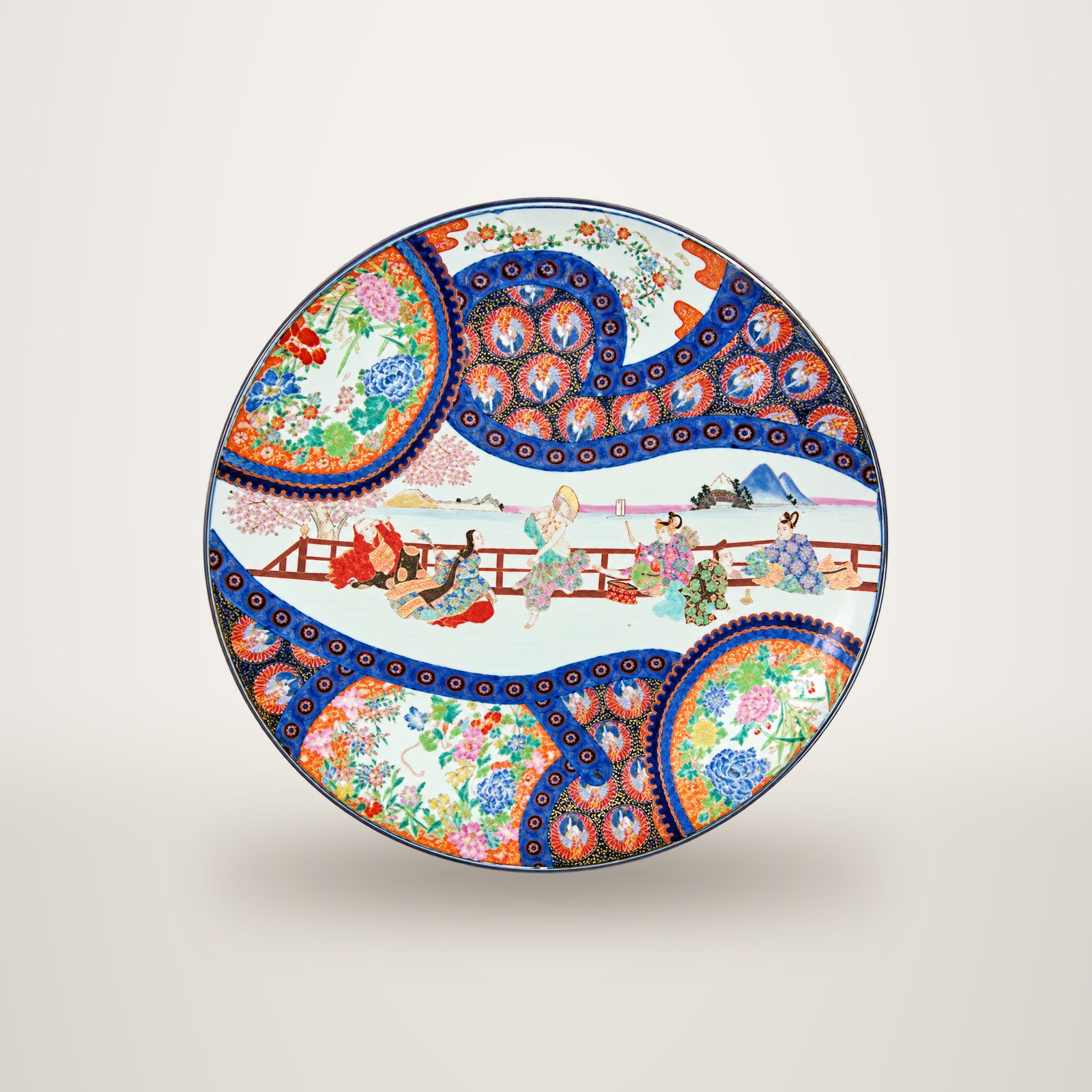 Grand plat rond en porcelaine aux motifs japonais colorés, avec une scène de personnages sur une terrasse et des médaillons de fleurs bleus et orange.