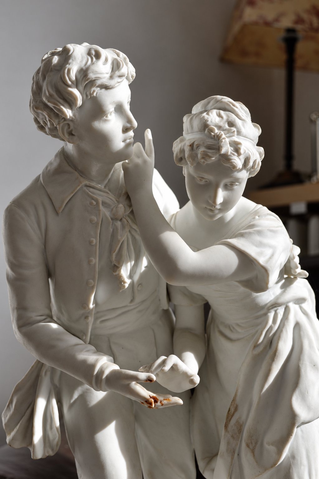 Sculpture en biscuit de porcelaine blanche représentant un jeune homme et une jeune femme, la main de celle-ci levée vers son menton, éclairée par une lumière rasante.