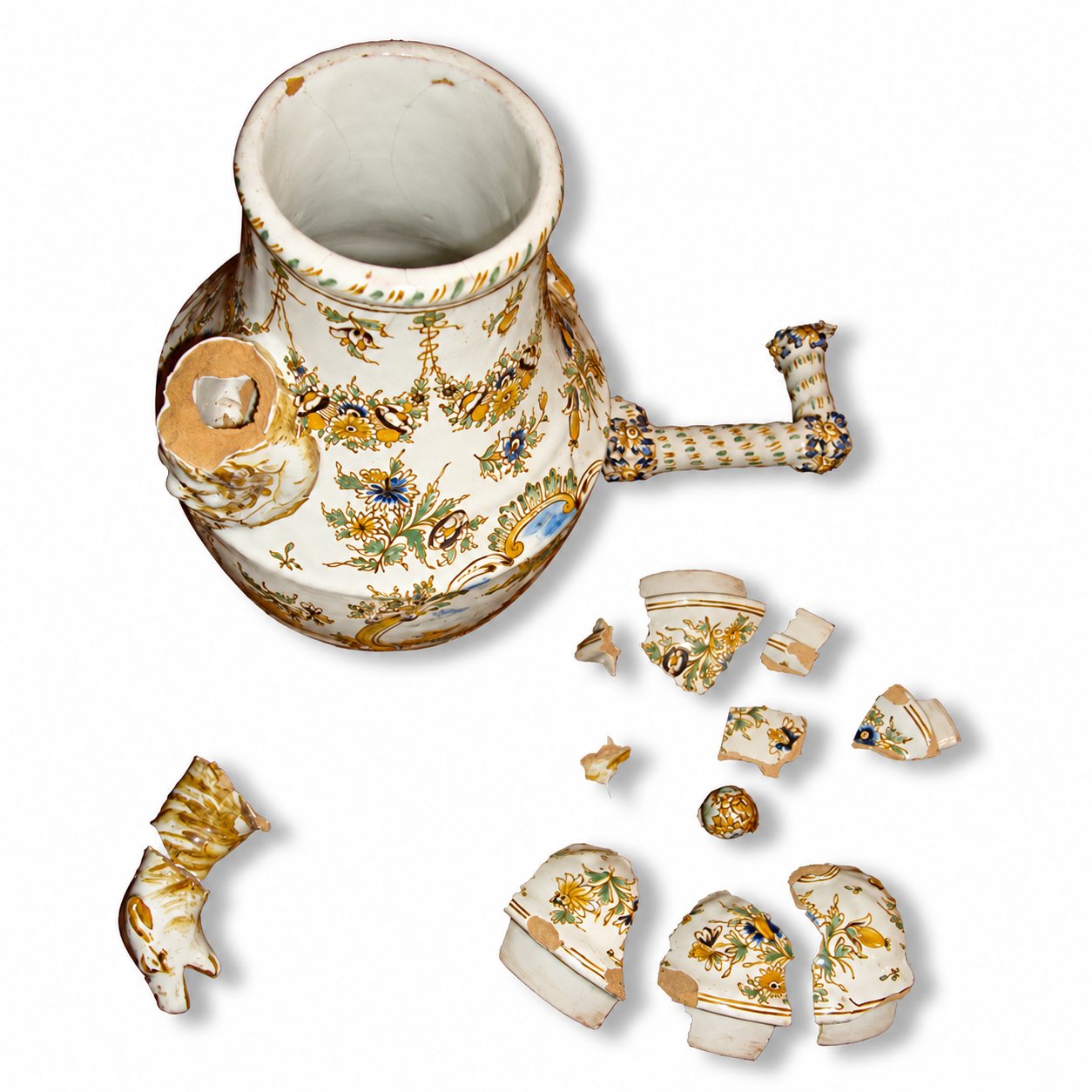 Un pichet en faïence à décor floral polychrome, dont l'anse est brisée, posé sur fond blanc à côté de nombreux éclats de céramique.