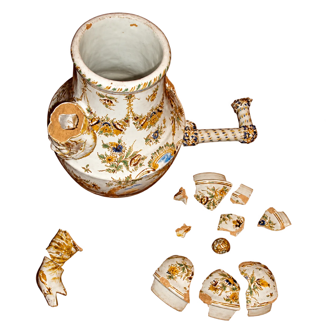 Un pichet en faïence à décor floral polychrome, cassé, posé sur fond blanc avec de nombreux morceaux détachés autour de lui.