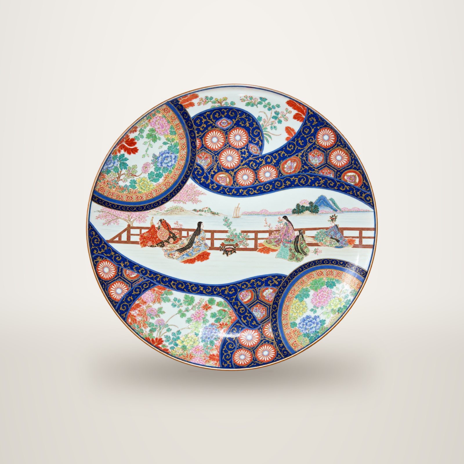 Un grand plat en porcelaine de style Imari, décoré de motifs floraux bleus, rouges et dorés et d'une scène de personnages près d'une barrière, photographié sur fond blanc.