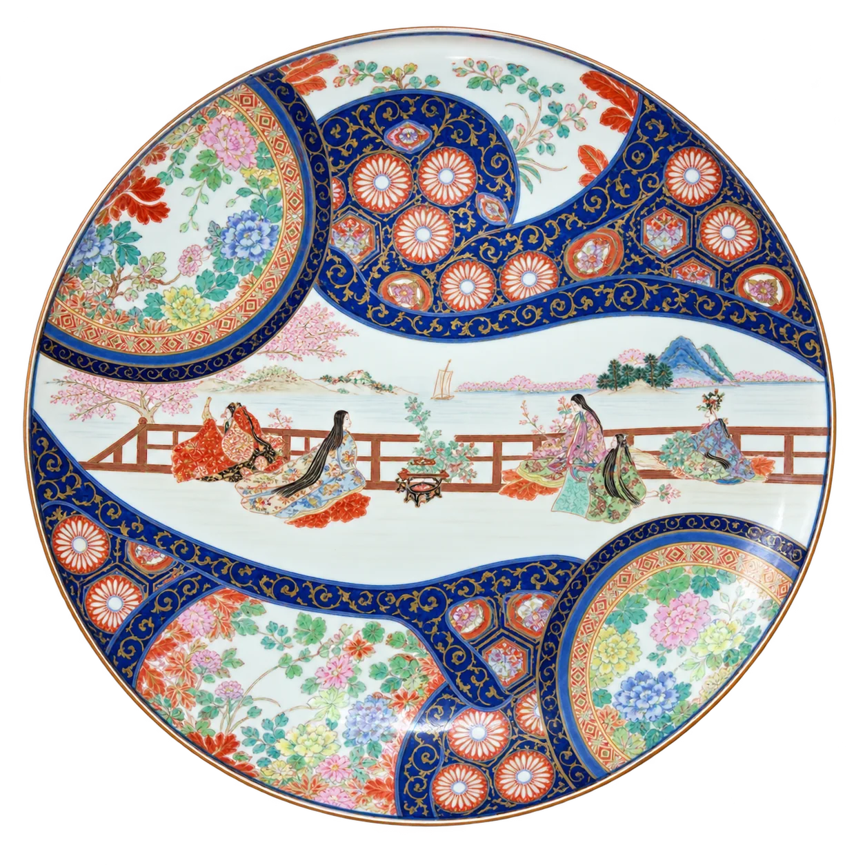 Un grand plat en porcelaine de style Imari, décoré de motifs floraux bleus, rouges et dorés et d'une scène de personnages près d'une barrière, photographié sur fond blanc.