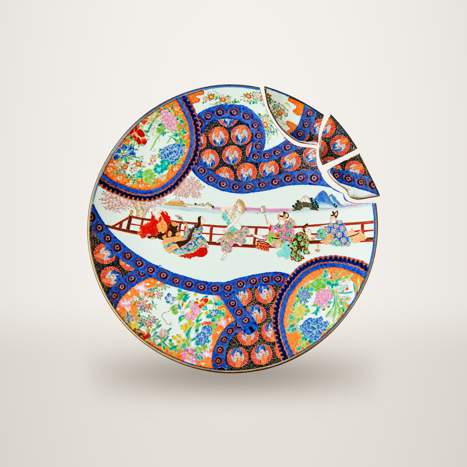 Grand plat en porcelaine aux motifs japonais colorés, bleu, orange et rouge, représentant des personnages près d'une balustrade, avec trois fragments détachés au bord supérieur droit.