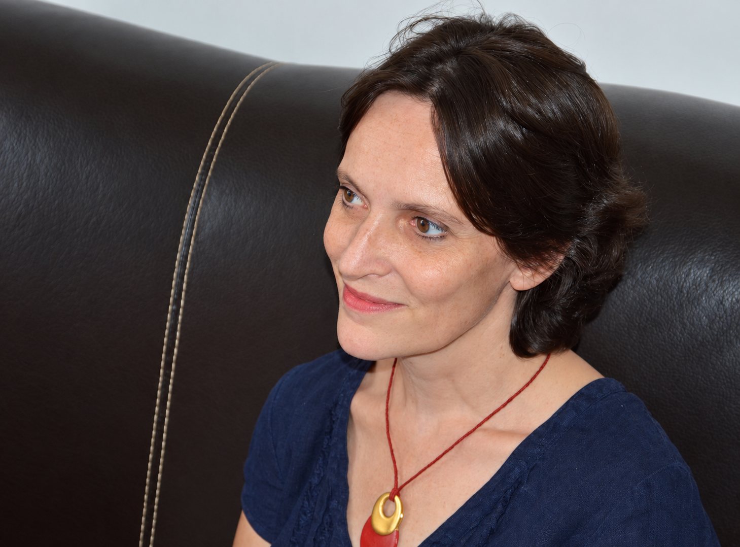 Une femme aux cheveux bruns attachés, vêtue d'un haut bleu marine et portant un pendentif doré sur un cordon rouge, sourit assise devant un canapé en cuir foncé.