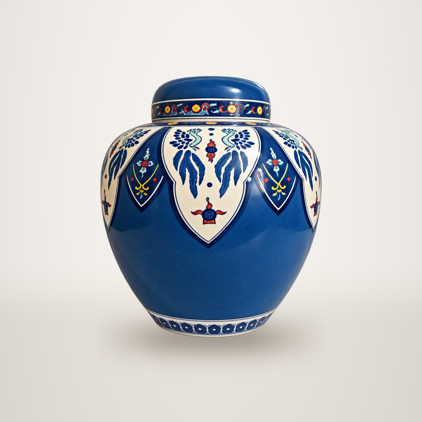 Un pot couvert en céramique bleue, orné de motifs floraux blancs, bleus et rouges, posé devant un fond uni blanc.