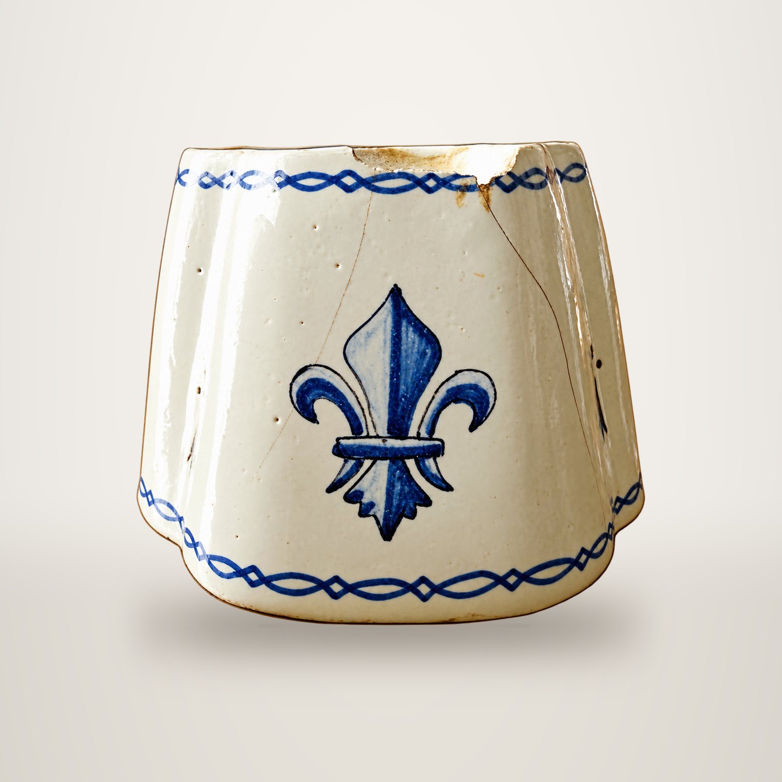 Un ancien pot en faïence blanche ébréché, orné d'une fleur de lys bleue et de frises bleues sur fond blanc.