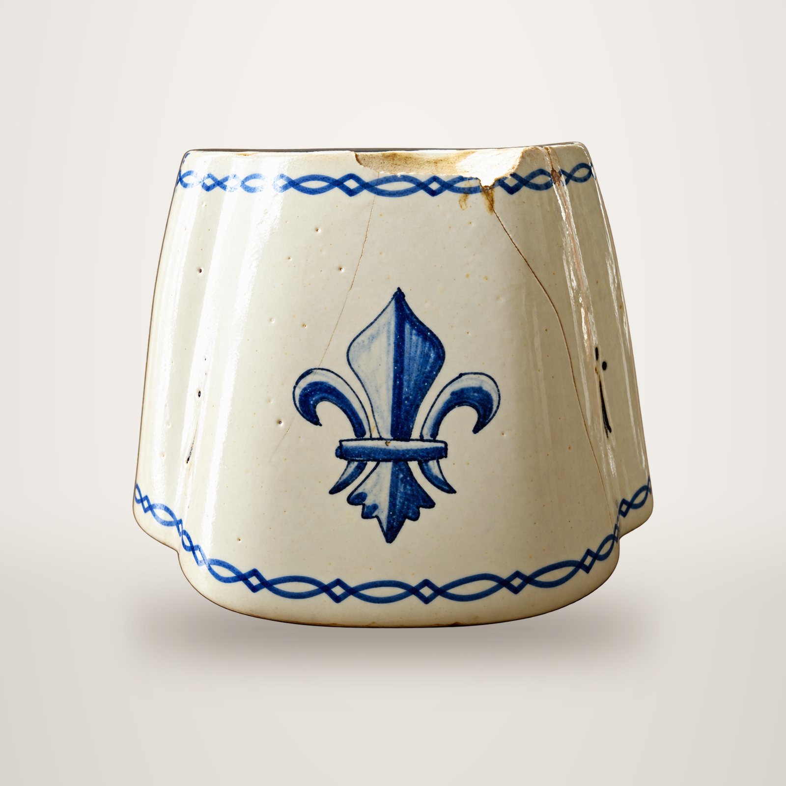 Un pot ancien en faïence blanche, orné d'une fleur de lys bleue et de frises bleues, présente quelques fissures et éclats.