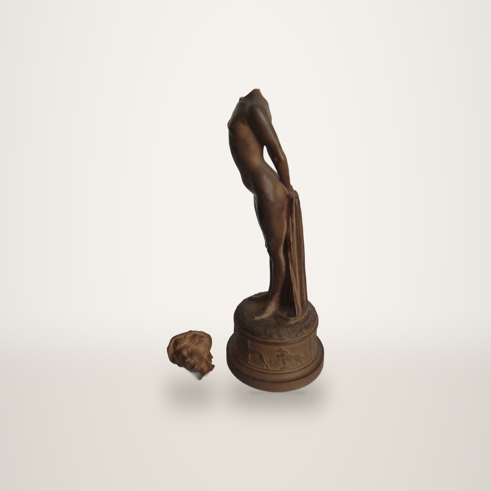 Une statuette en terre cuite brune représentant une femme nue, dont la tête cassée est posée à côté du socle orné d'animaux en relief.