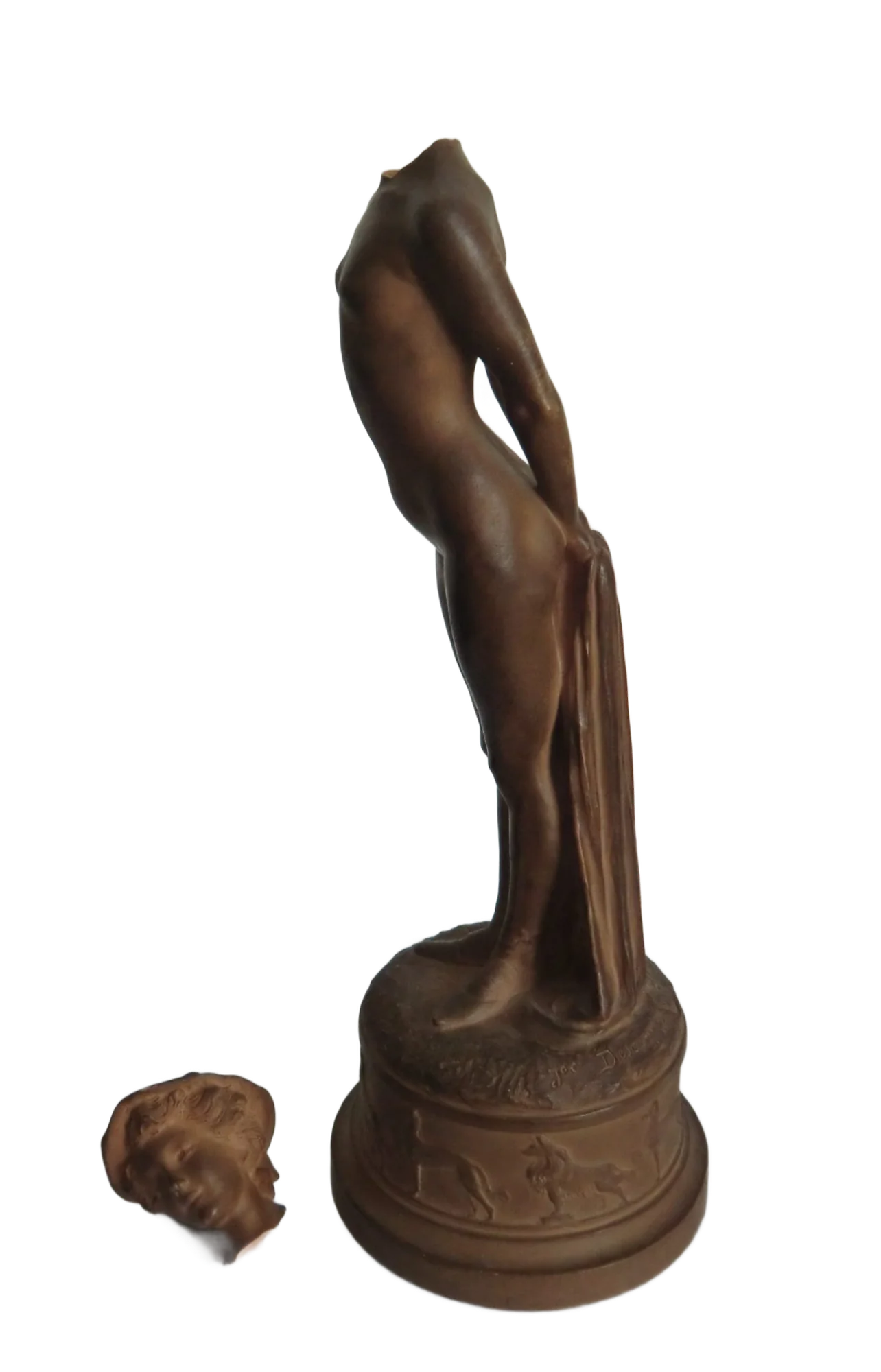 Une statuette en terre cuite brune représentant une femme nue, dont la tête cassée est posée à côté du socle orné d'animaux en relief.