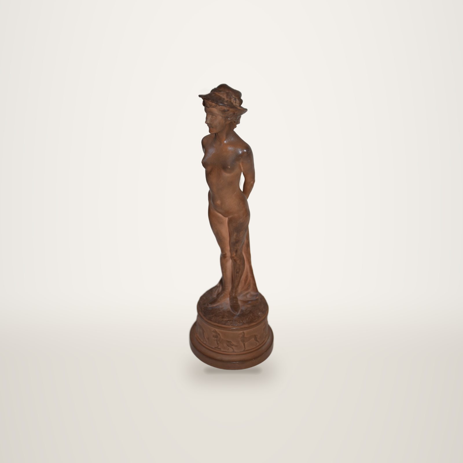 Statuette en terre cuite représentant une femme nue coiffée d'un chapeau, debout sur un socle rond orné de chiens en relief, devant un mur blanc.