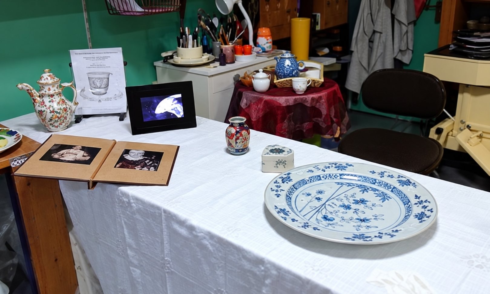 Une table recouverte d'une nappe blanche présente une grande assiette en porcelaine bleue et blanche, un album ouvert, un petit cadre et des objets anciens, avec d'autres bibelots en arrière-plan.