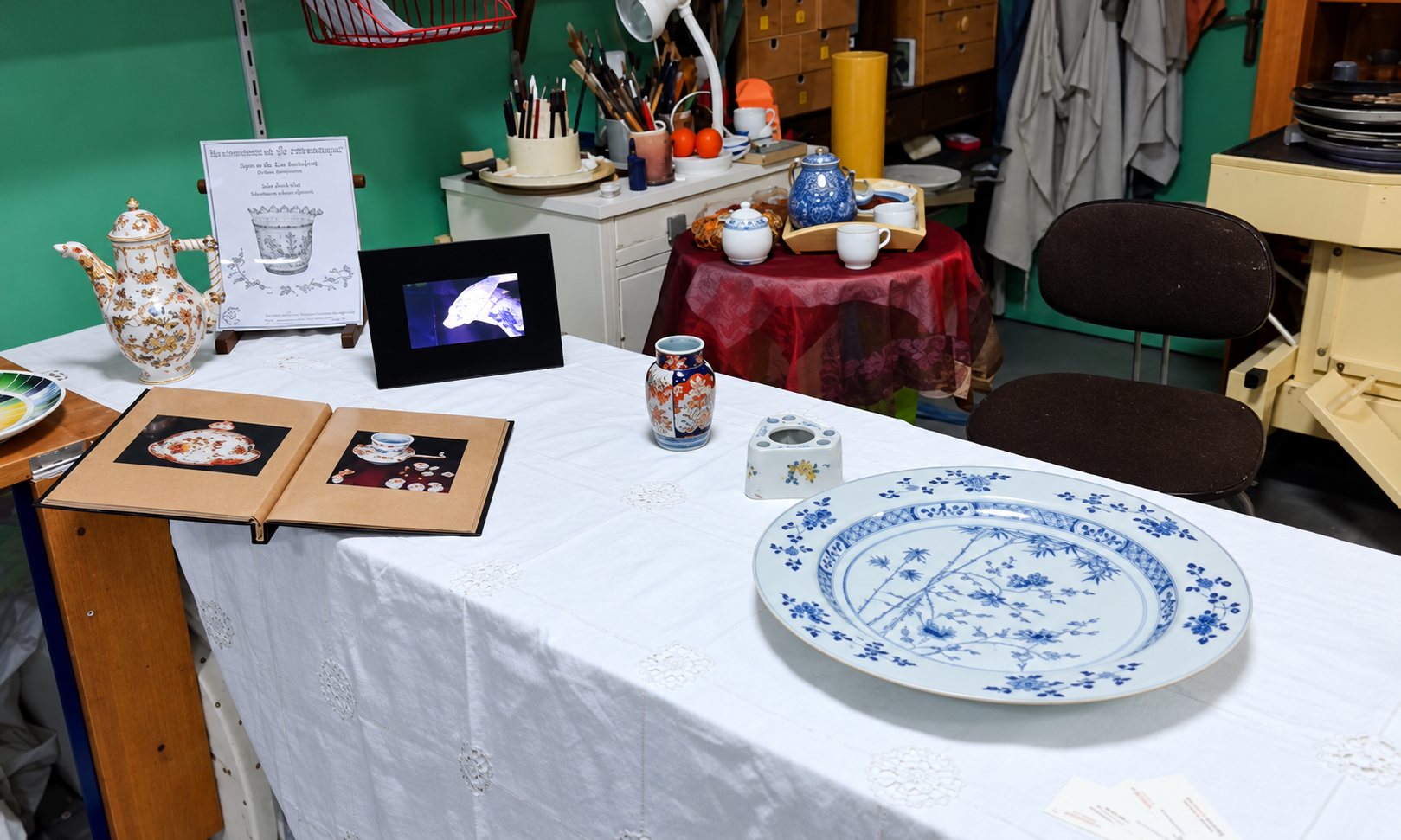 Une table recouverte d'une nappe blanche présente une grande assiette en porcelaine bleue et blanche, une théière décorée, un petit vase, un album photo ouvert et un cadre numérique dans un atelier.