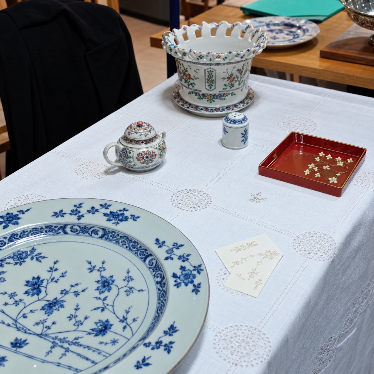 Une table recouverte d'une nappe blanche présente une grande assiette décorée de motifs bleus, une théière, un compotier ajouré et un petit plateau rouge.