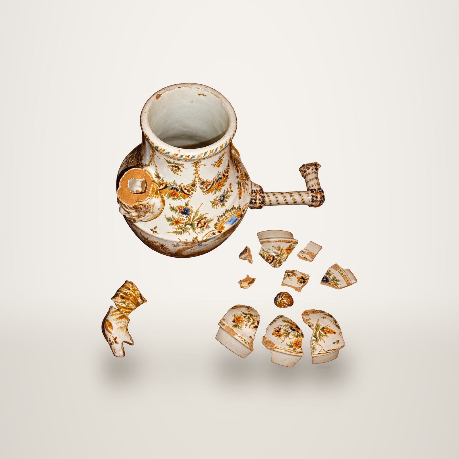 Un vase ancien en faïence à décor floral peint, cassé, posé sur fond blanc avec son anse torsadée détachée et de nombreux fragments autour.